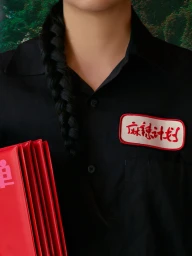 Close-up portrait of a person in a black collared shirt with a red and white embroidered patch, holding a bright red folder.
