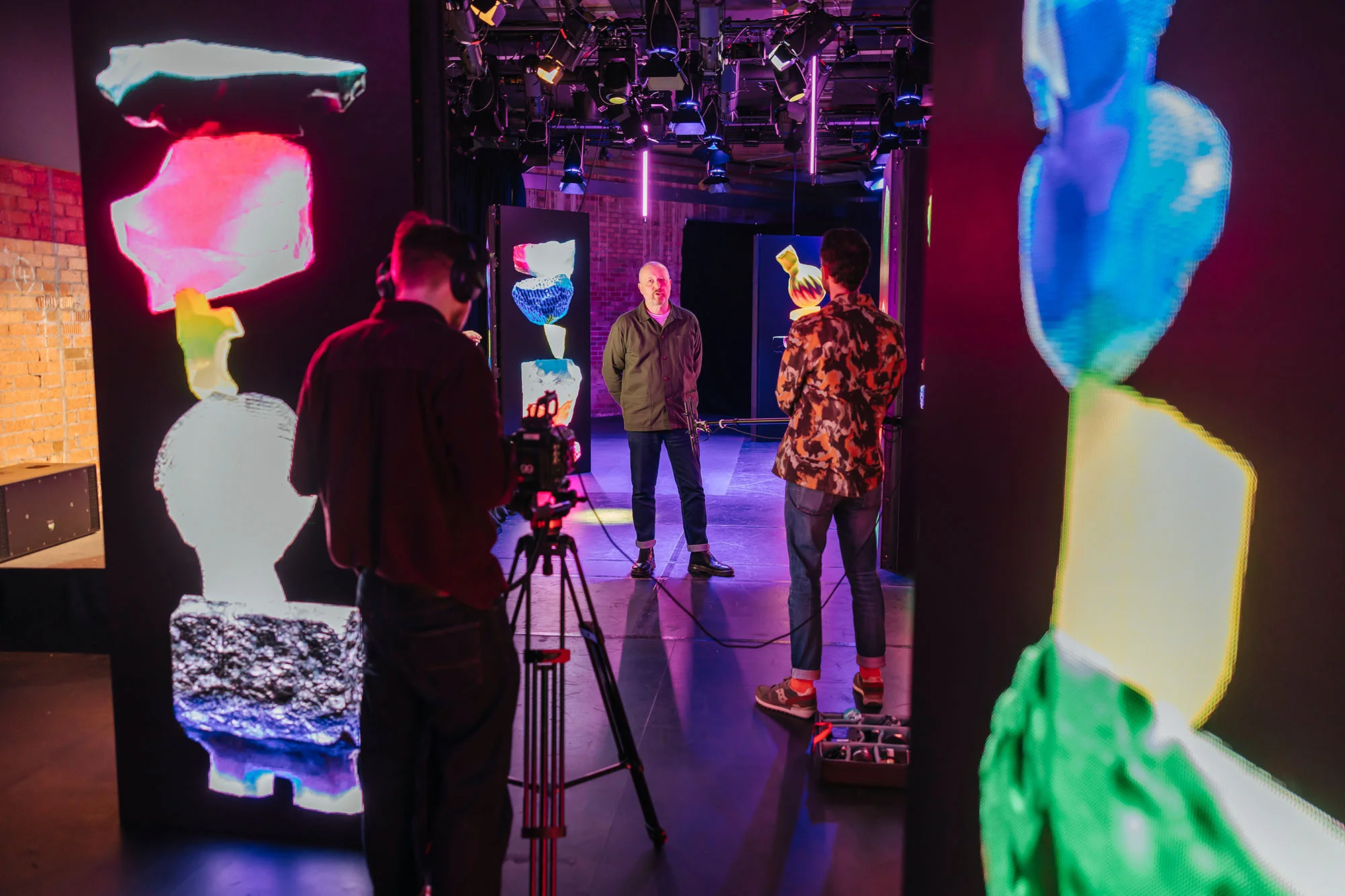 On a photo set, a man stands illuminated between two cameramen and abstract, colorful LED panels in an indoor photo studio environment.