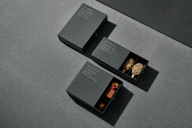 Overhead shot of three matte black Pana Chocolate boxes on a gray textured surface, with focus on the chocolates inside the open boxes.