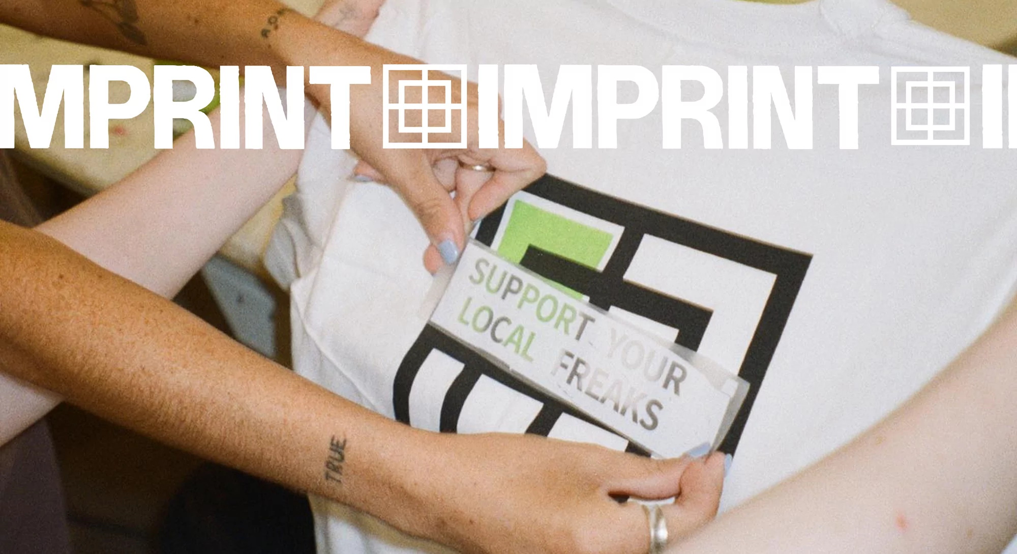 Close-up of a white t-shirt being customized with a black and green logo by two sets of hands with tattoos, promoting local artists.