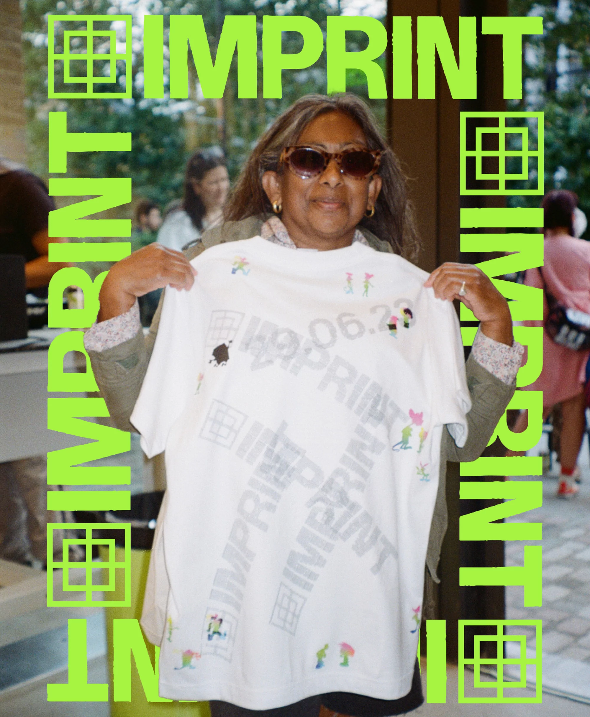 An older woman wearing sunglasses holds up a white t-shirt with graphic designs in an outdoor environment with light-green framing elements.