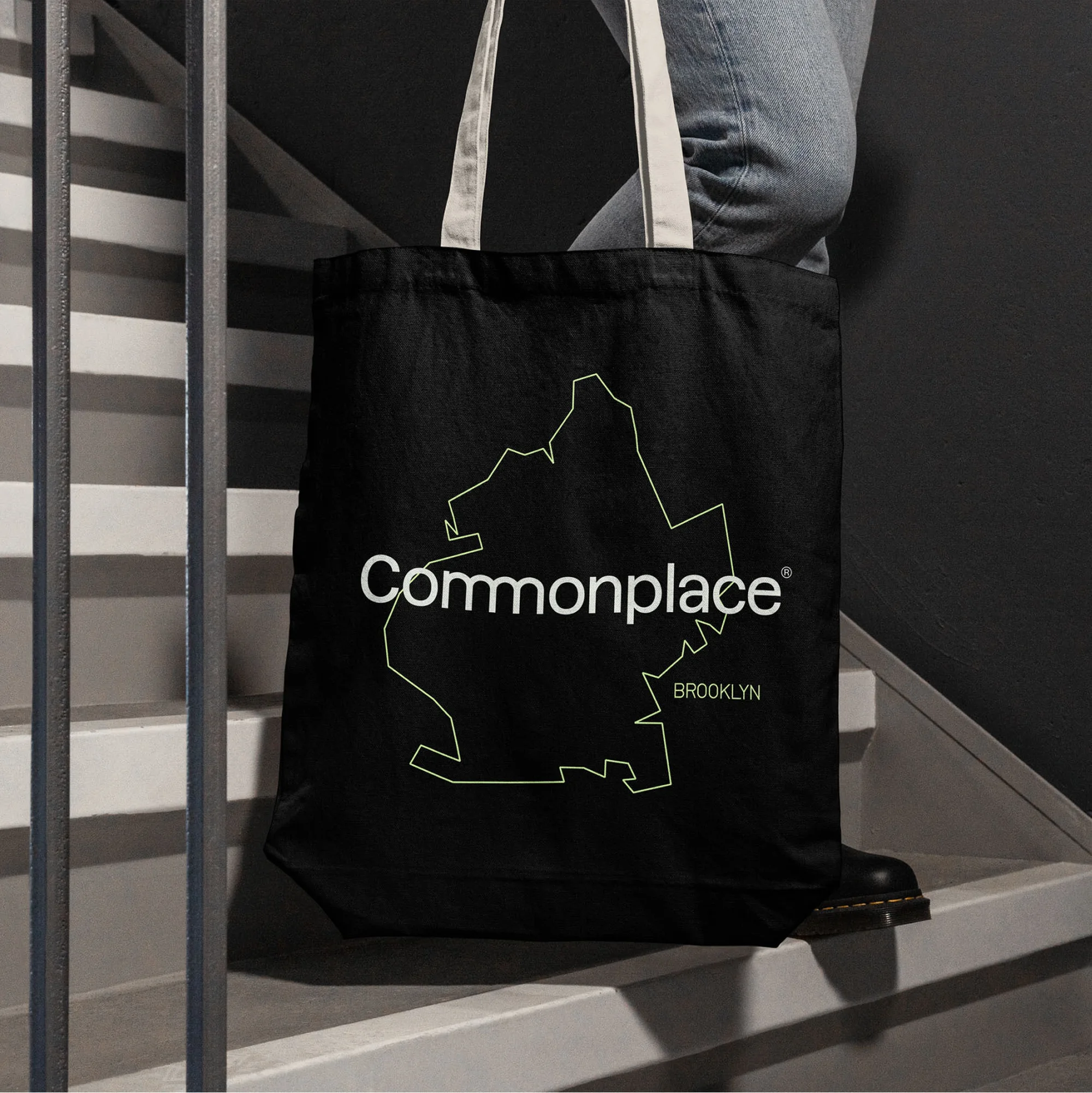 Person stands on stairs holding a black tote bag with the text 'Commonplace BROOKLYN' and a Brooklyn map graphic.