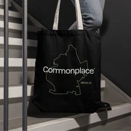 Black canvas tote bag with white Commonplace brand logo held by a person walking down concrete stairs