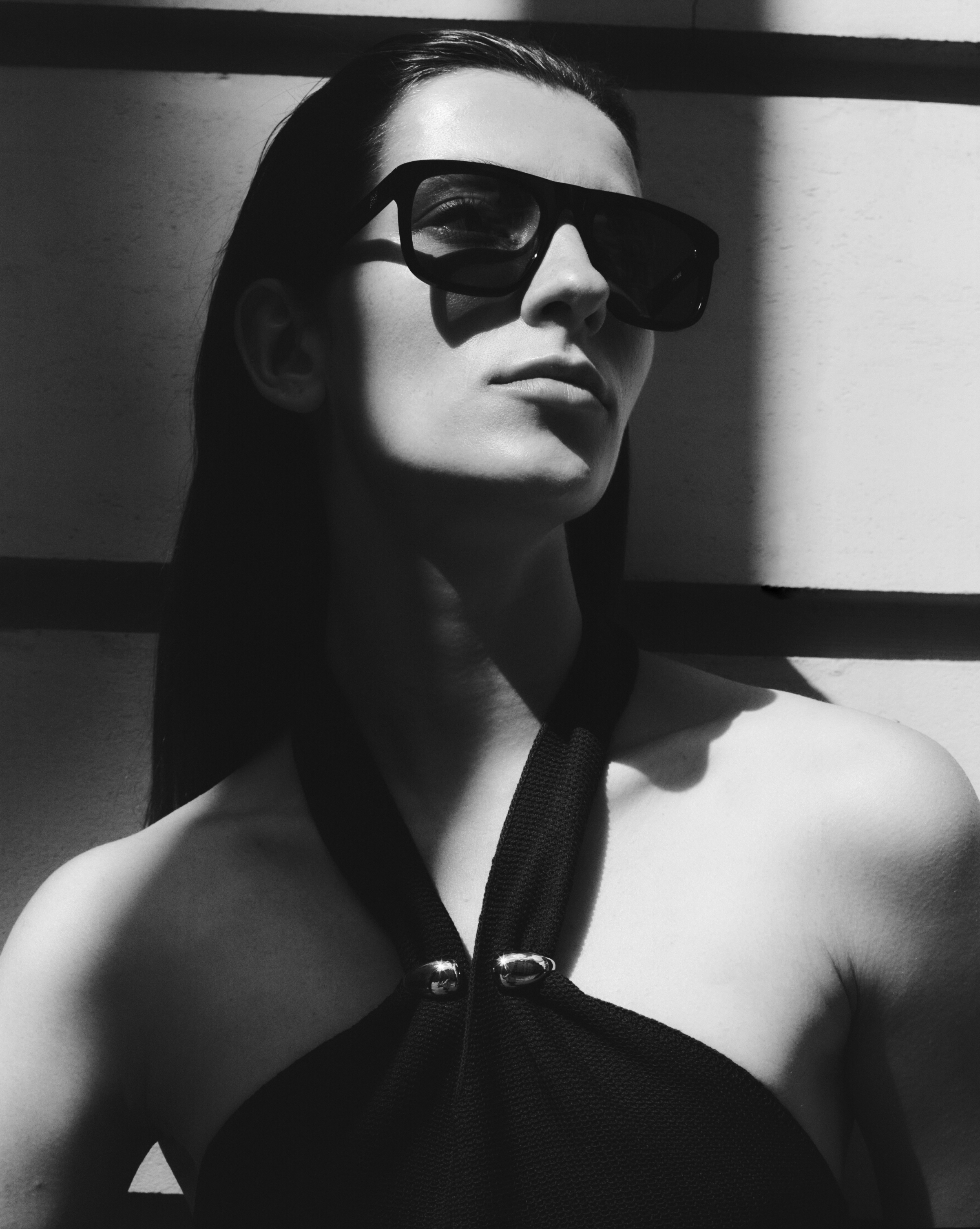 Black and white portrait of a woman with dark hair wearing dark sunglasses and a black top, standing in front of a wall with light and shadow.