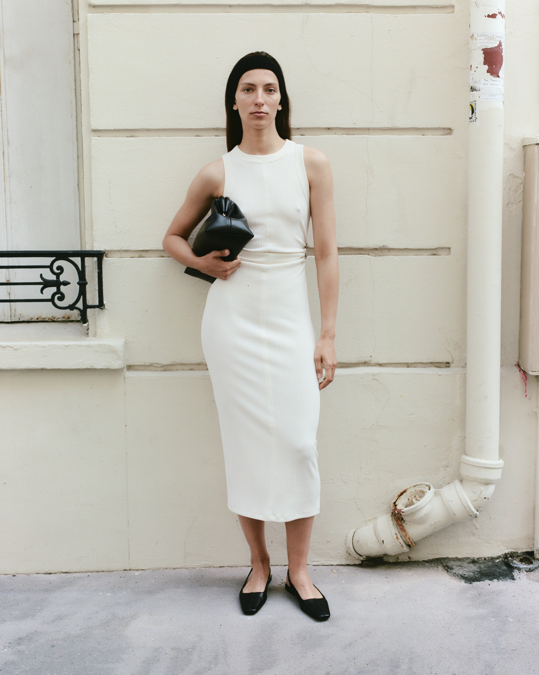 A slender woman poses in a cream-colored dress with a black clutch and black flats in an outdoor setting with a neutral background.