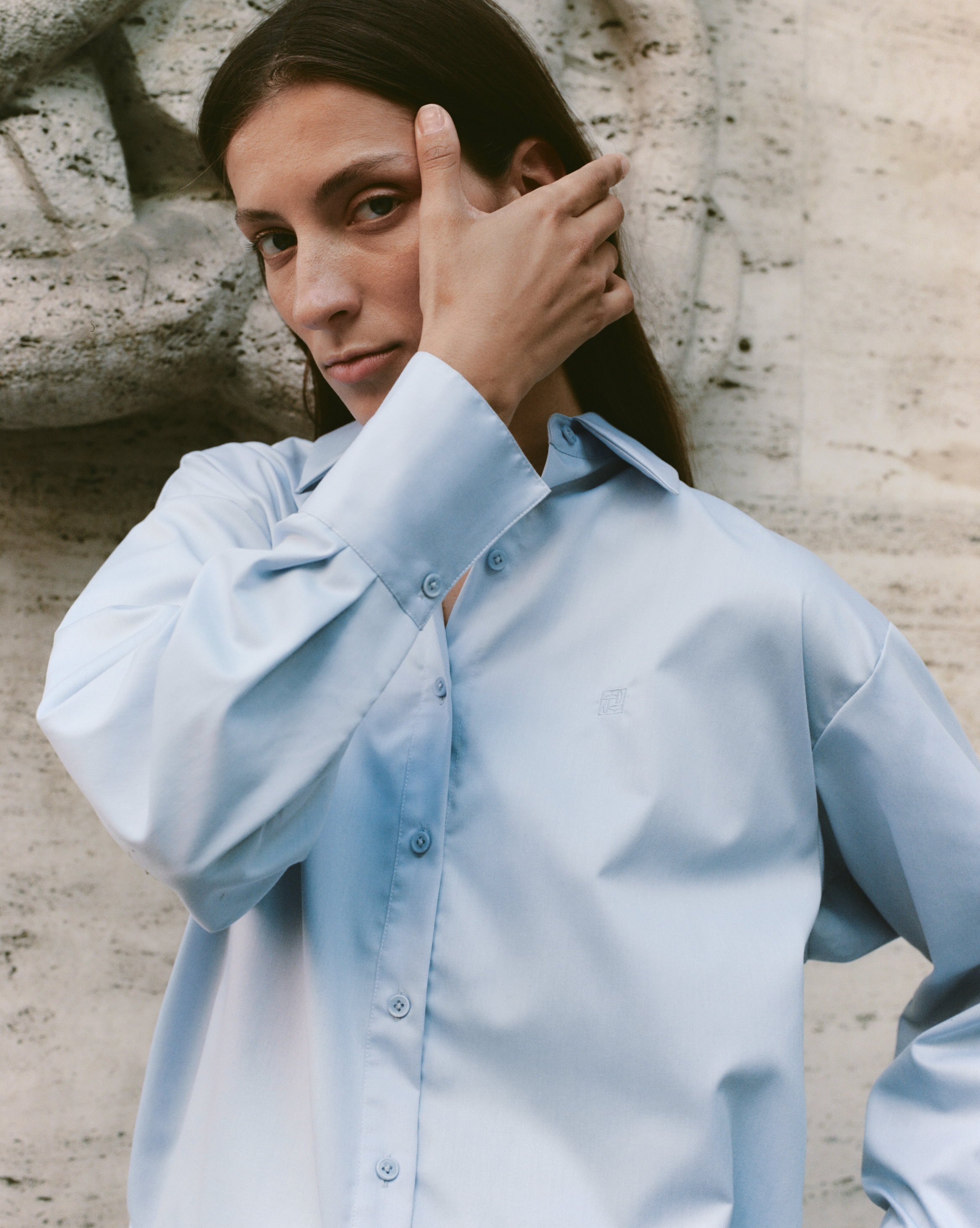 A brunette woman with long hair wears a light blue button-down shirt against a textured wall, offering a contemplative, direct gaze.
