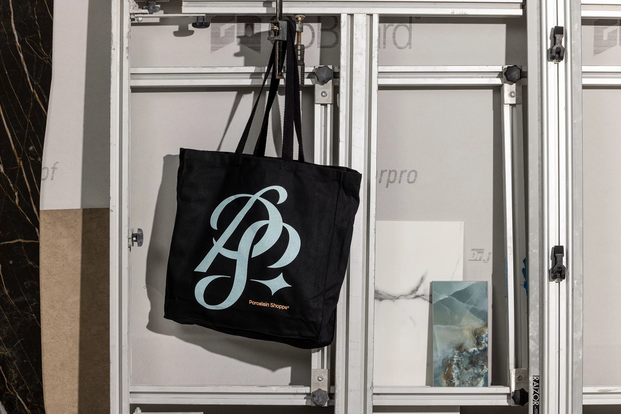 Black tote bag with a white logo, hanging in a bright indoor setting with marble and porcelain tile samples on a display rack.