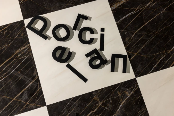 Overhead view of black letter cutouts spelling 'Porcelain' on a floor with black and white marble tiles, arranged on a white tile.