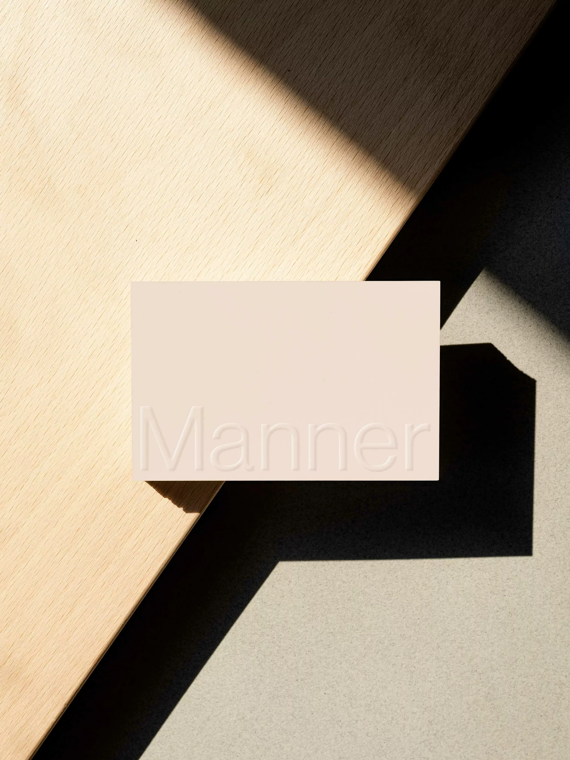 Beige business card with debossed Manner logo resting on wooden and stone surfaces casting diagonal shadows.