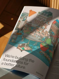 An open magazine spread featuring an isometric architectural illustration with buildings, tiny figures, and typography reading We are building the foundations for a better future.