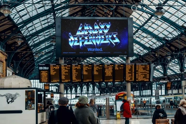Large digital advertising screen displaying metallic futuristic typography inside a busy Victorian railway station concourse.