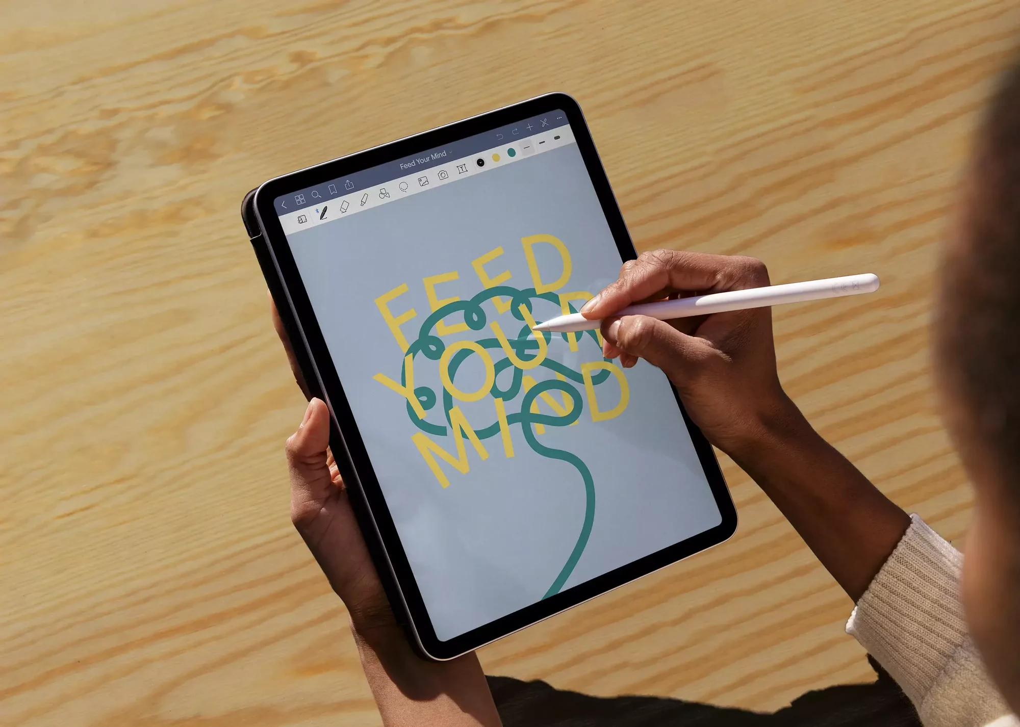 Hands hold tablet with digital artwork of brain and the text 'Feed Your Mind.' Person draws on the screen with a white stylus pen.