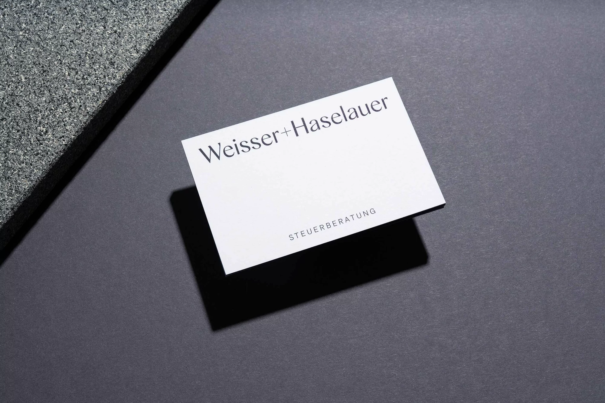 Clean, minimalist product shot featuring a white business card with black text on a dark gray surface, paired with textured gray stone.