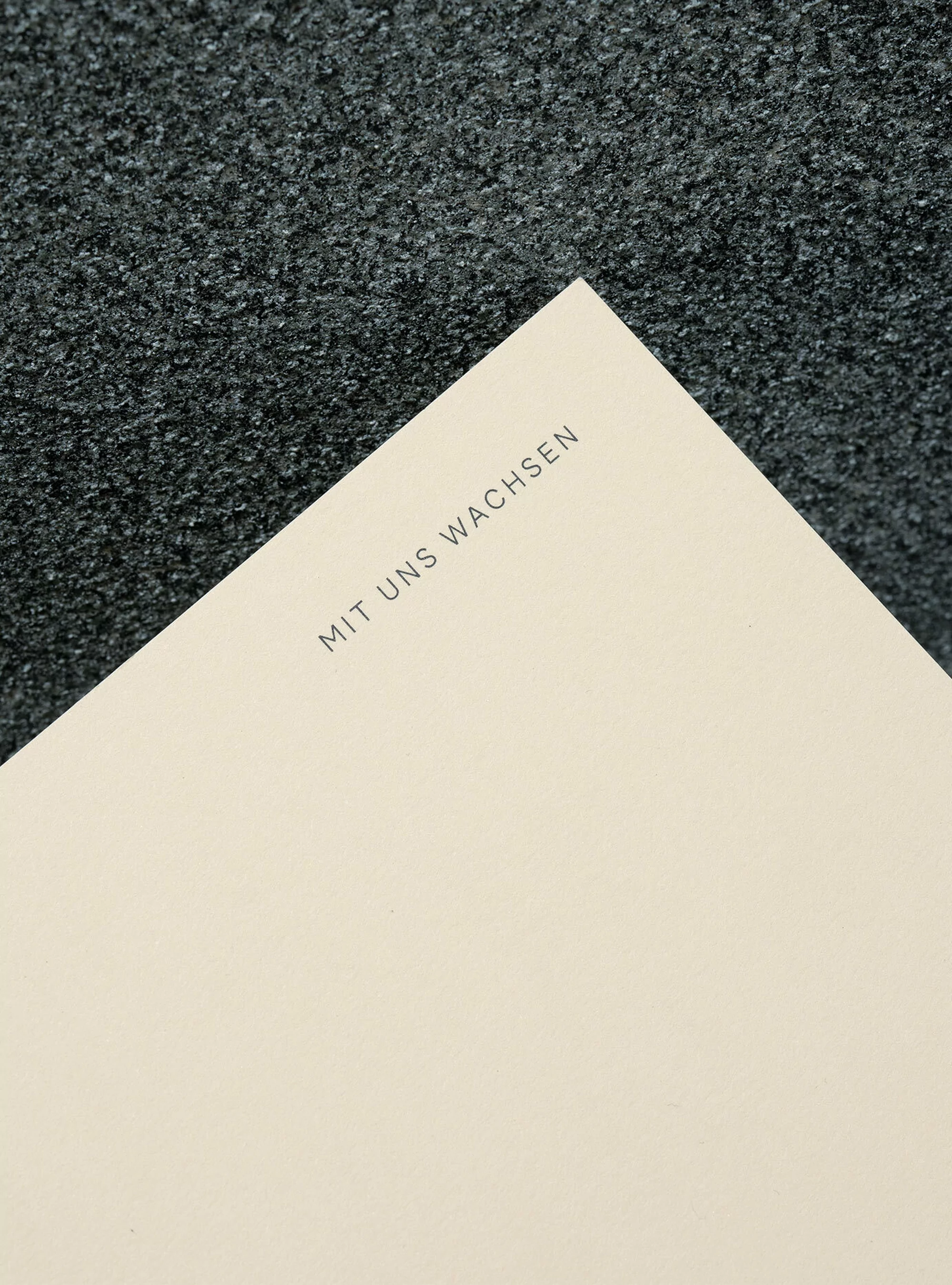 Minimalist image of a beige paper corner with the German text 'Mit Uns Wachsen' against a textured, dark grey background.