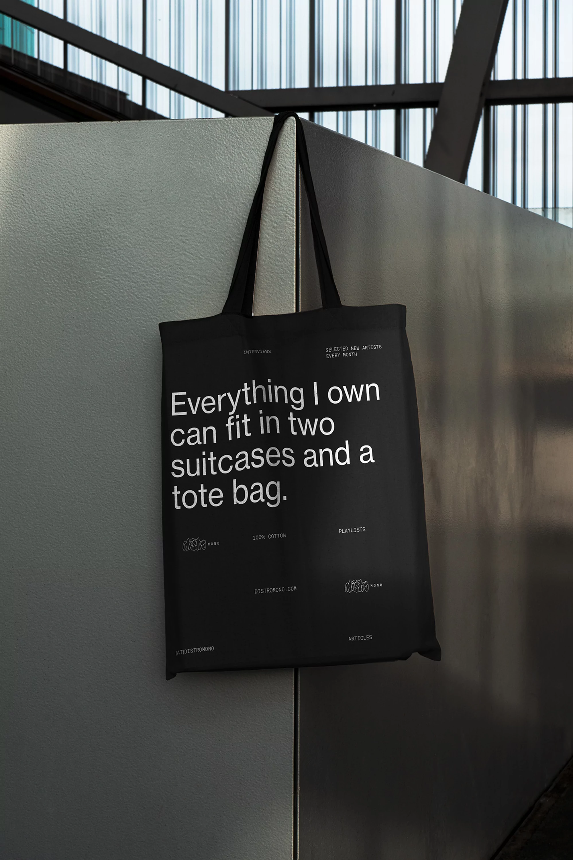 Black tote bag hanging against a gray wall with a metal framework ceiling. Features a printed white quote and logo designs.