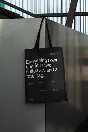 Black tote bag hanging against a gray wall with a metal framework ceiling. Features a printed white quote and logo designs.