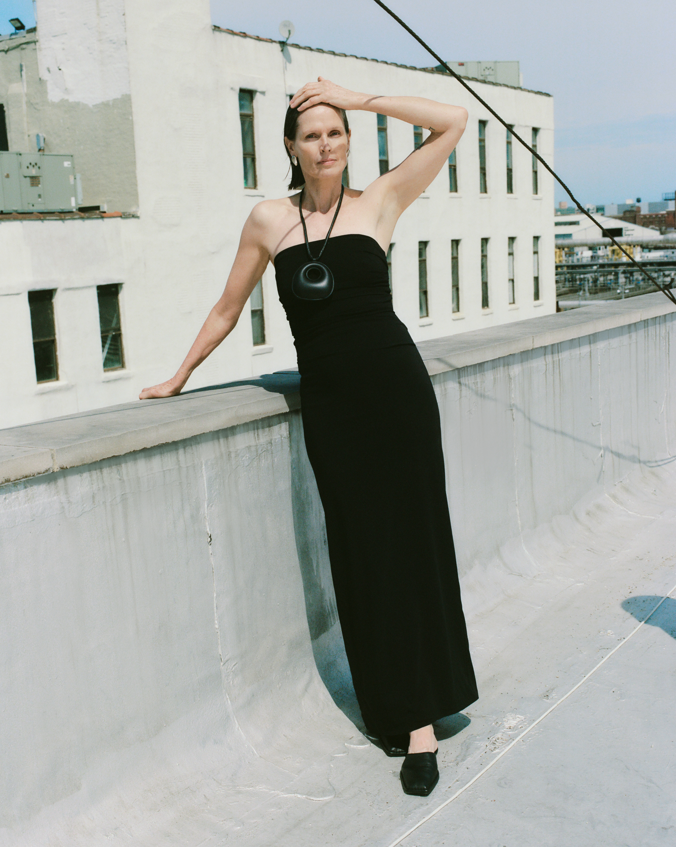 Elegant woman in a black dress and pendant poses on a rooftop with city buildings in the background under a bright, sunny sky.