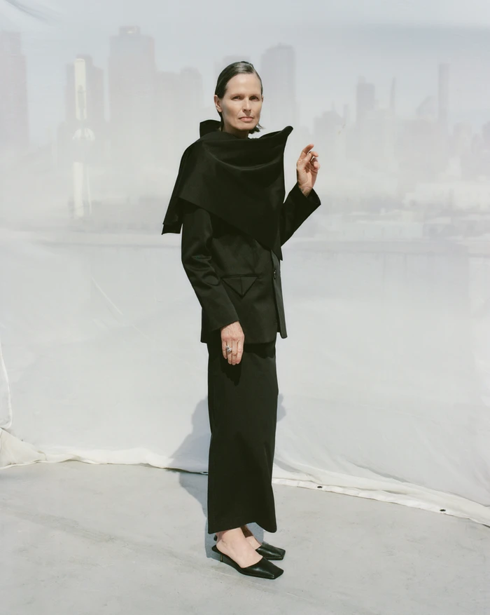 Model in a black structured blazer and long skirt posing against a draped white backdrop for St Agni.