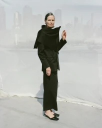 Model in a black structured blazer and long skirt posing against a draped white backdrop for St Agni.
