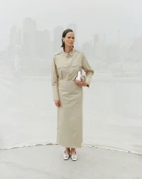 Model standing outdoors wearing a beige shirt and matching skirt holding a small silver bag with city skyline in background