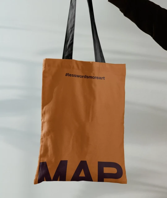 Earth-toned tote bag displaying '#lesswordsmoreart' and 'MAP' texts against a light gray backdrop held by someone with dark clothing.