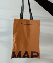 Tan tote bag with black handles and black lettering held against a light wall