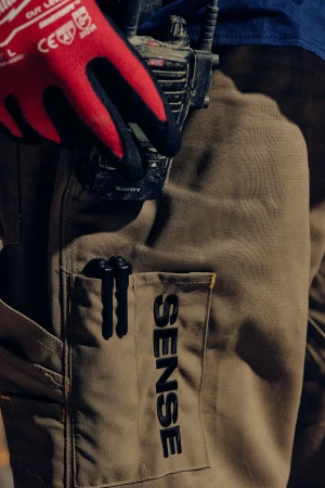 Close-up of a construction worker's tan work pants featuring a pocket with the word 'SENSE' and a red-gloved hand holding a walkie-talkie.