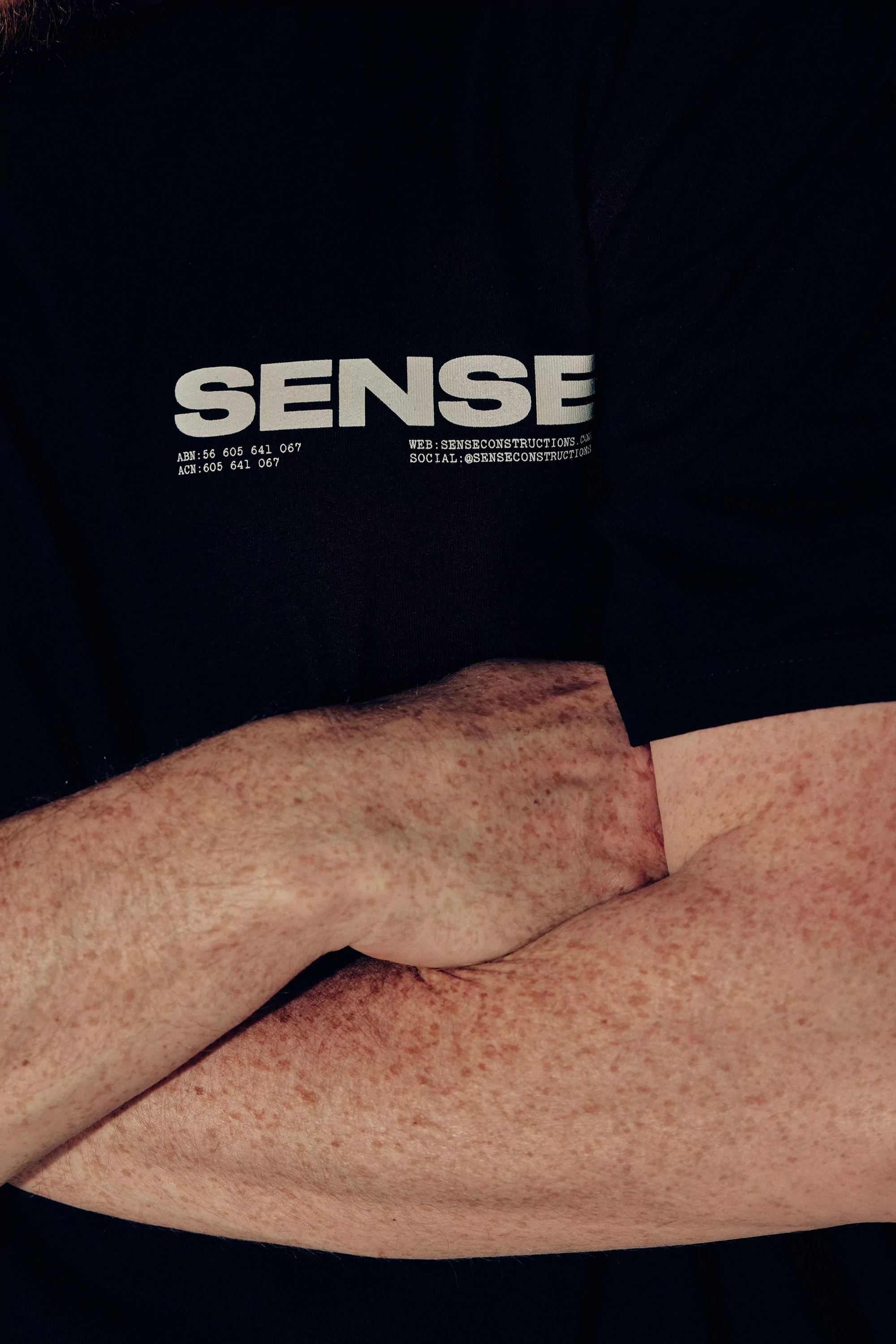Close-up of fair-skinned arms crossing in front of a black t-shirt with the word SENSE printed in white, demonstrating a minimalist style.