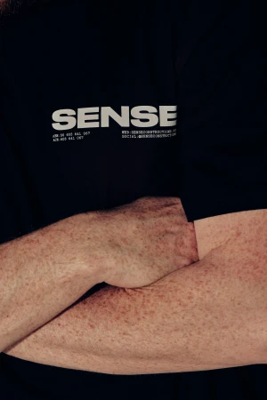 Close-up of fair-skinned arms crossing in front of a black t-shirt with the word SENSE printed in white, demonstrating a minimalist style.