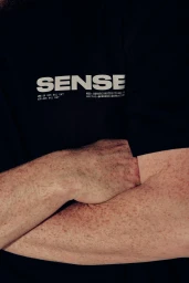 Close-up of a person with freckled arms crossed wearing a black t-shirt with white Sense Constructions logo.