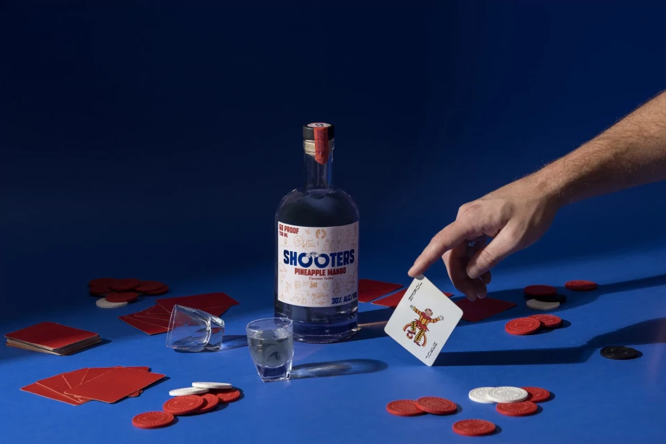 Bottle of Shooters pineapple mango flavored vodka with playing cards, shot glass, and poker chips on a blue surface.