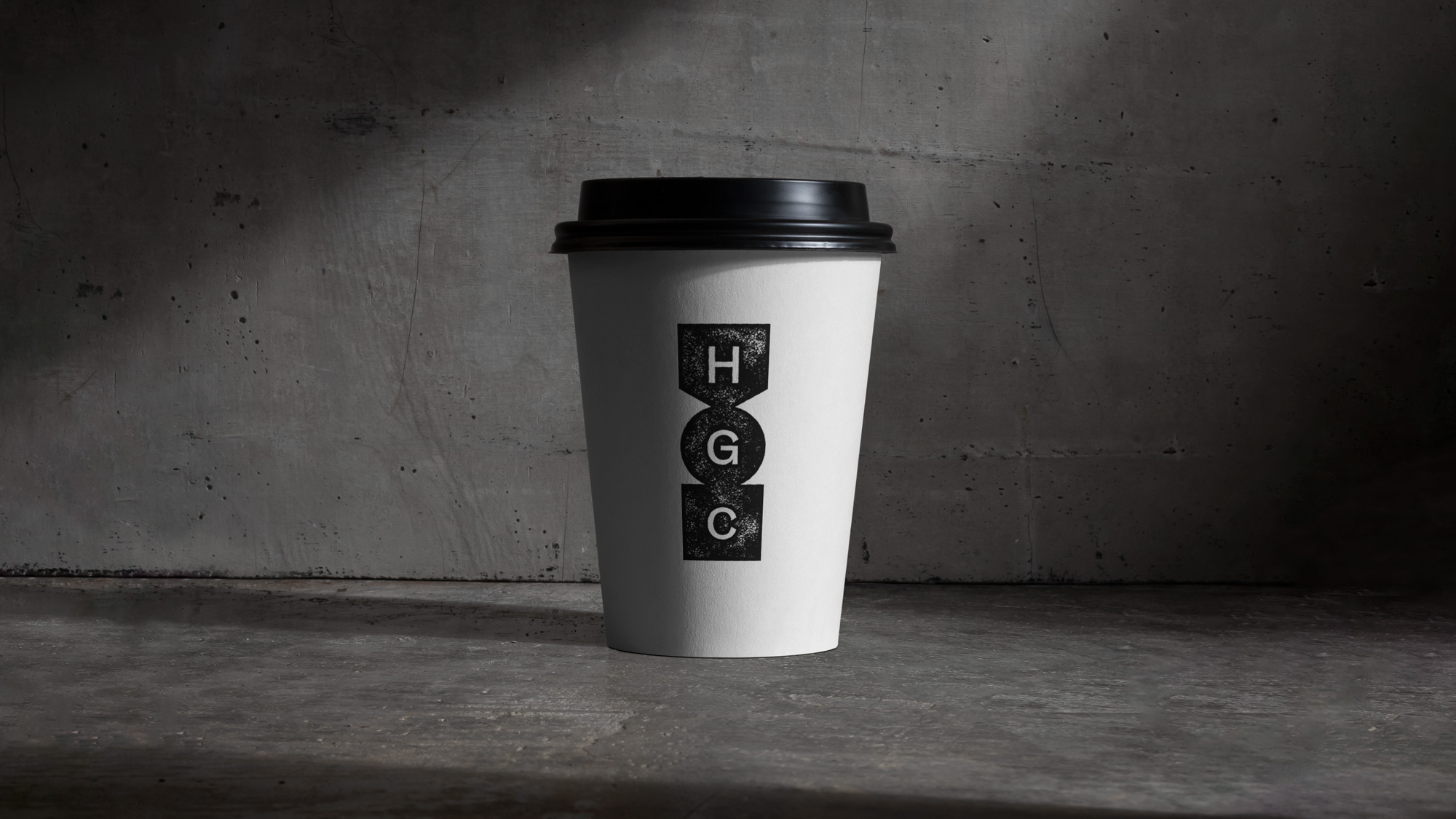 Paper coffee cup with black lid and HGC logo on a concrete background. Simple product shot with minimalist style and neutral colors.