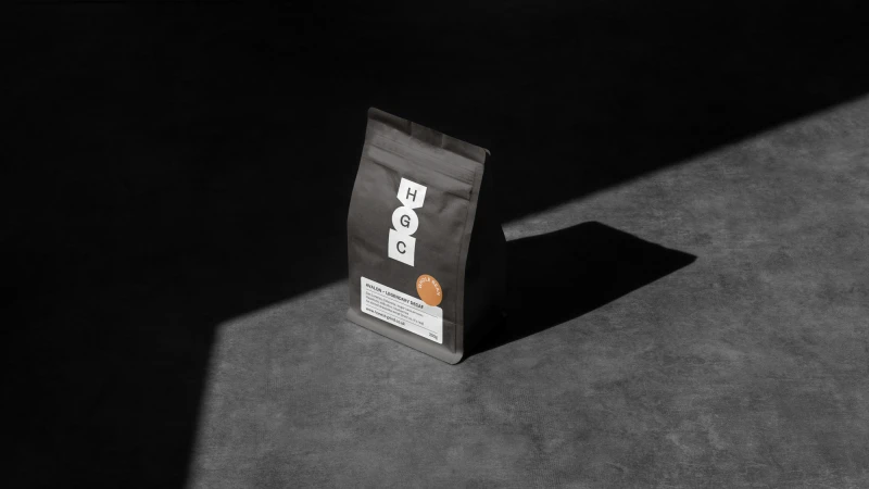 Minimalist studio shot of a black paper bag with white and orange branding, placed on a dark gray surface with strong shadows.