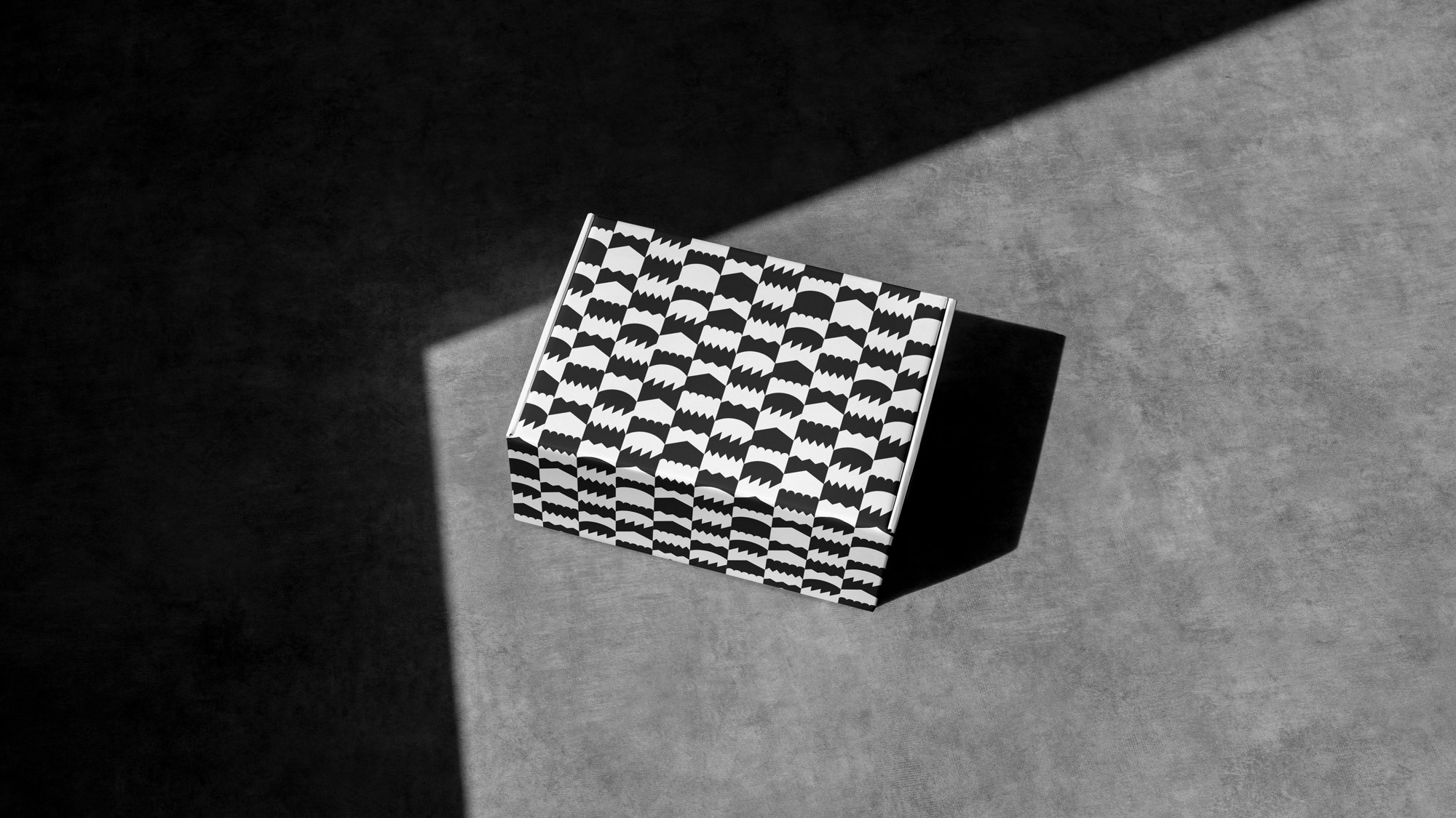 Monochromatic shot of a black-and-white patterned box on a concrete surface illuminated by a ray of light casting a shadow on the background.