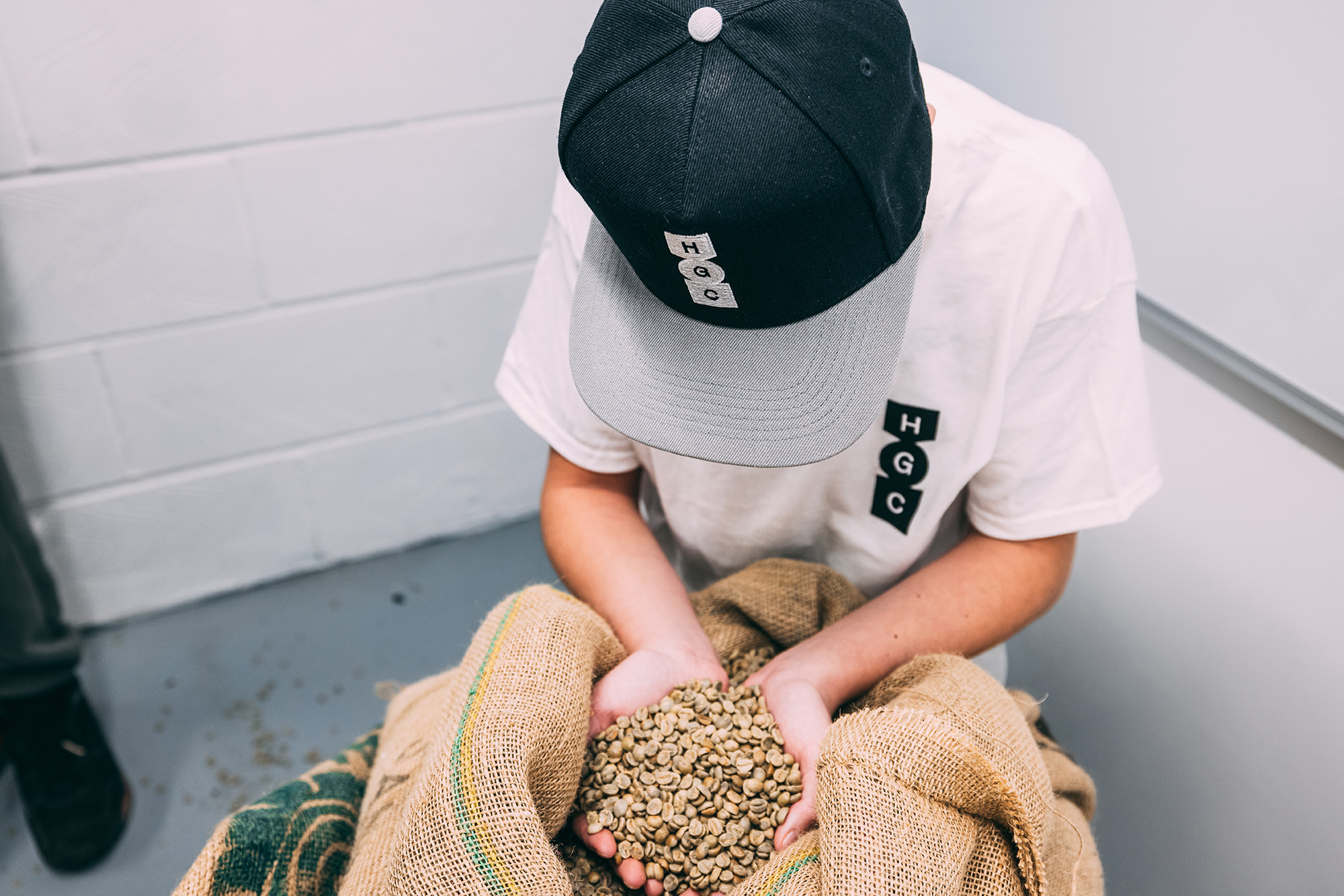 Person in a baseball cap holds green coffee beans in their hands over a burlap sack inside of a coffee production facility.