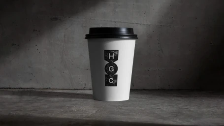 A white takeaway coffee cup with a black lid and stacked HGC logo stands on concrete against a textured wall.