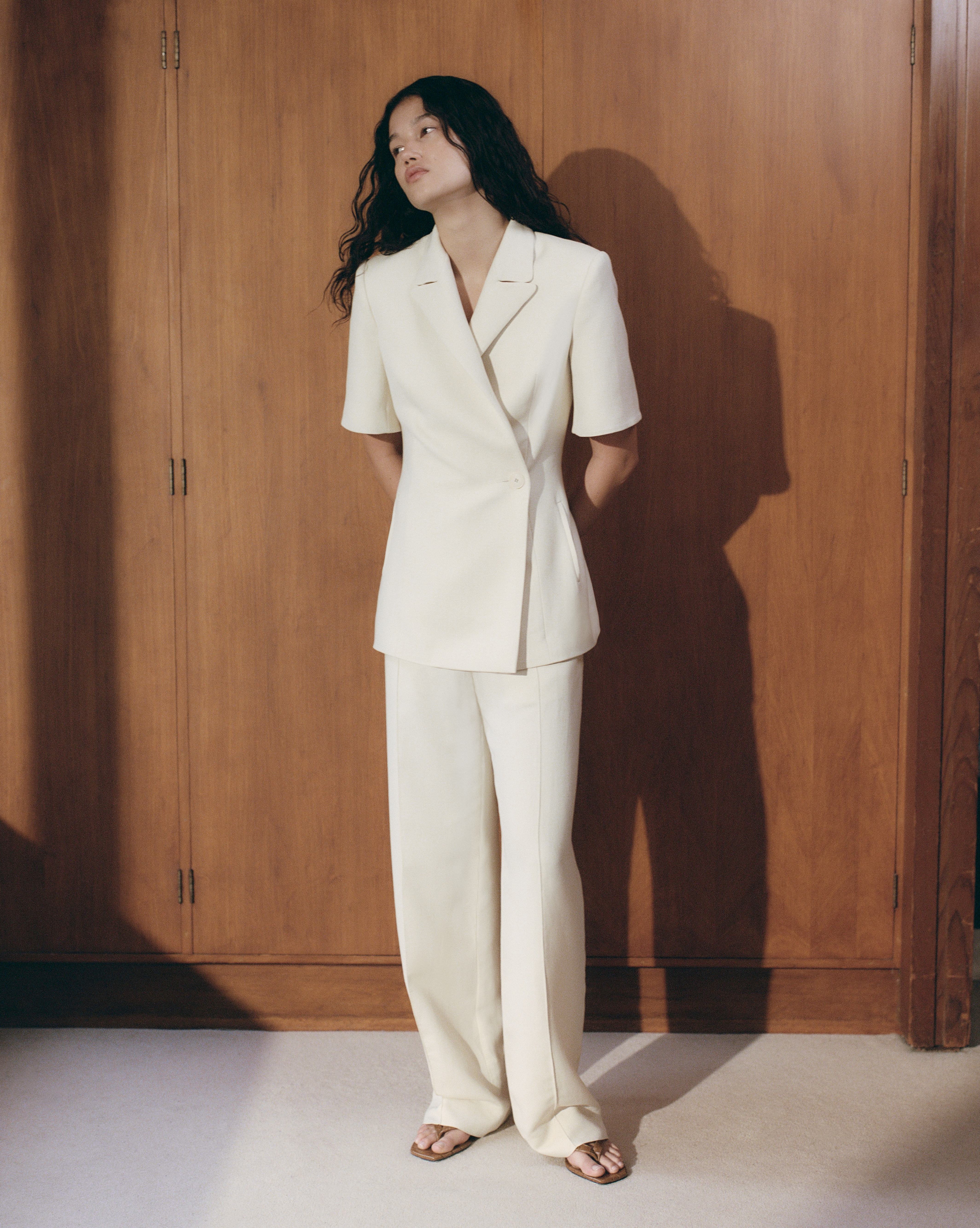 A full shot of a woman in an off-white suit, standing against a wood wall, wearing sandals. She has long dark hair and a neutral expression.