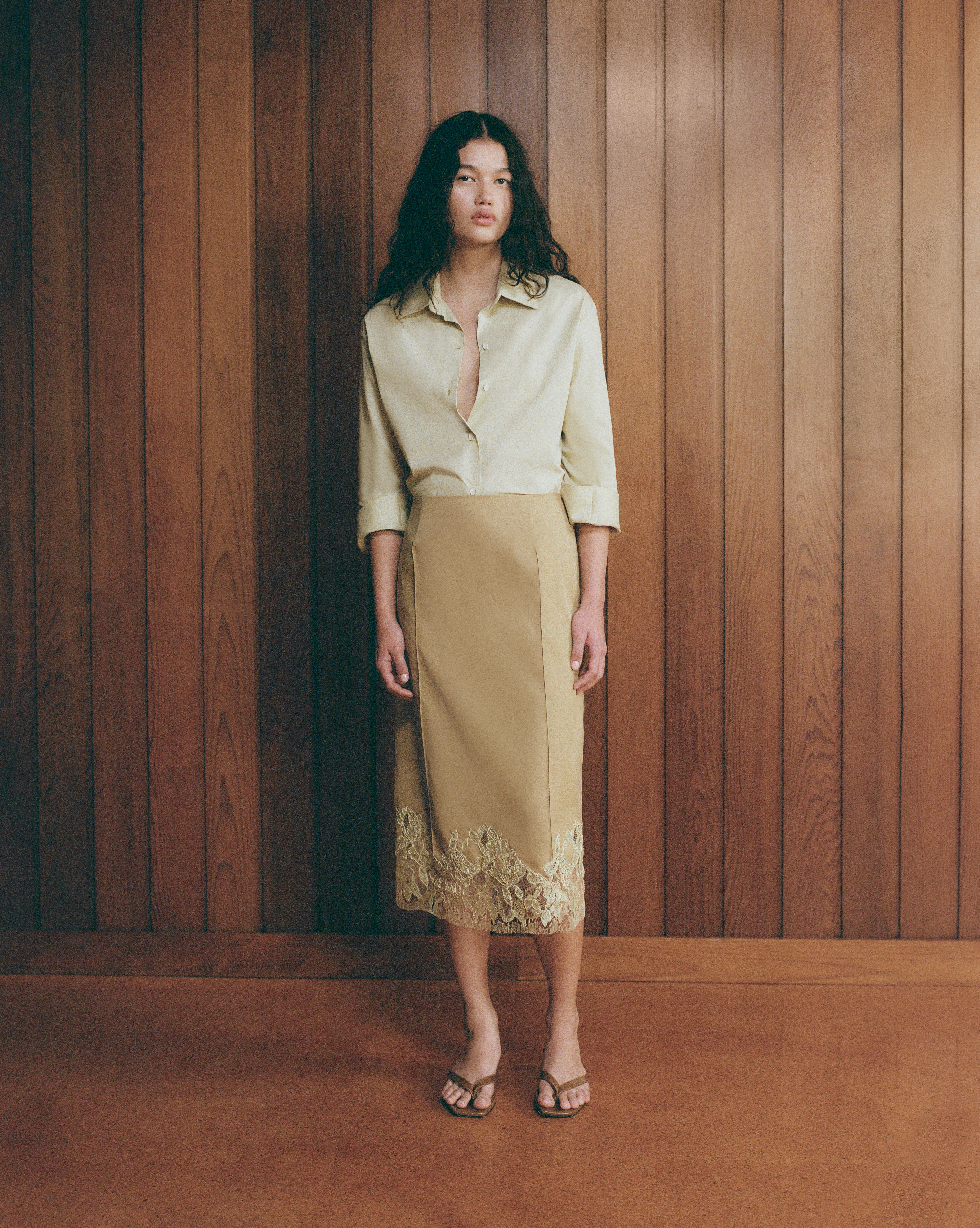 A young woman poses in a beige skirt with lace trim and a pale-yellow unbuttoned blouse. She stands against a wooden-paneled wall.