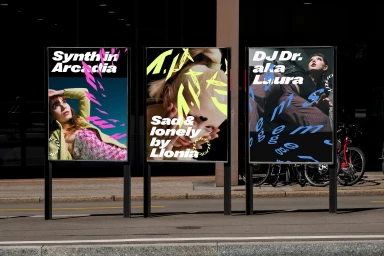 Three vertical promotional posters displaying graphic design and typography mounted outdoors along a city sidewalk.