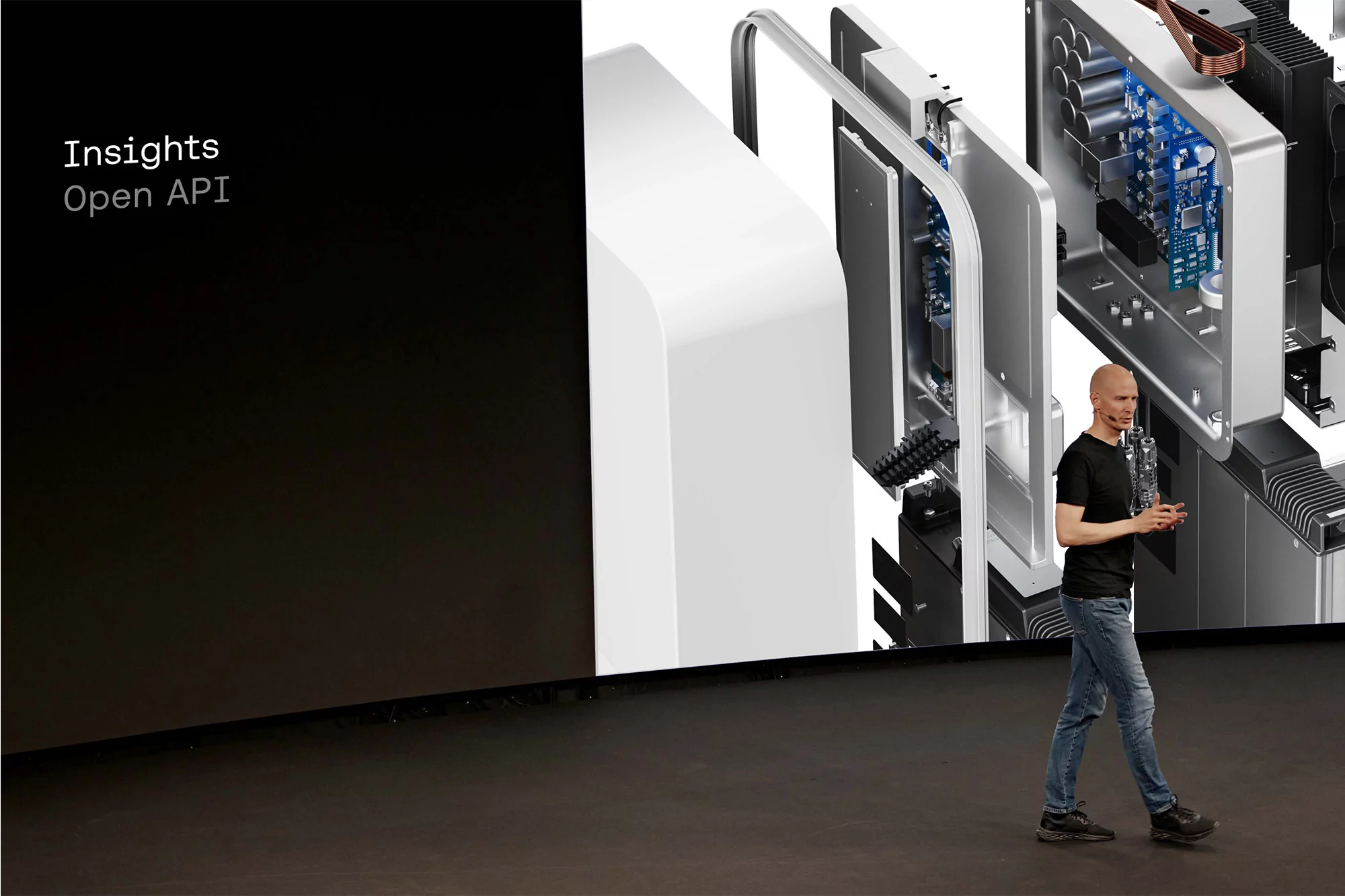 Adult male walks on stage during a tech presentation, with computer components displayed in the background, under bright stage lighting.
