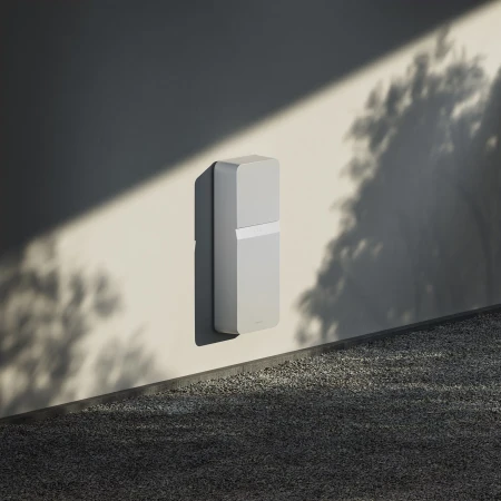 Modern matte-white rectangular electrical box mounted on a textured wall next to a gravel surface, outside with filtered sunlight.