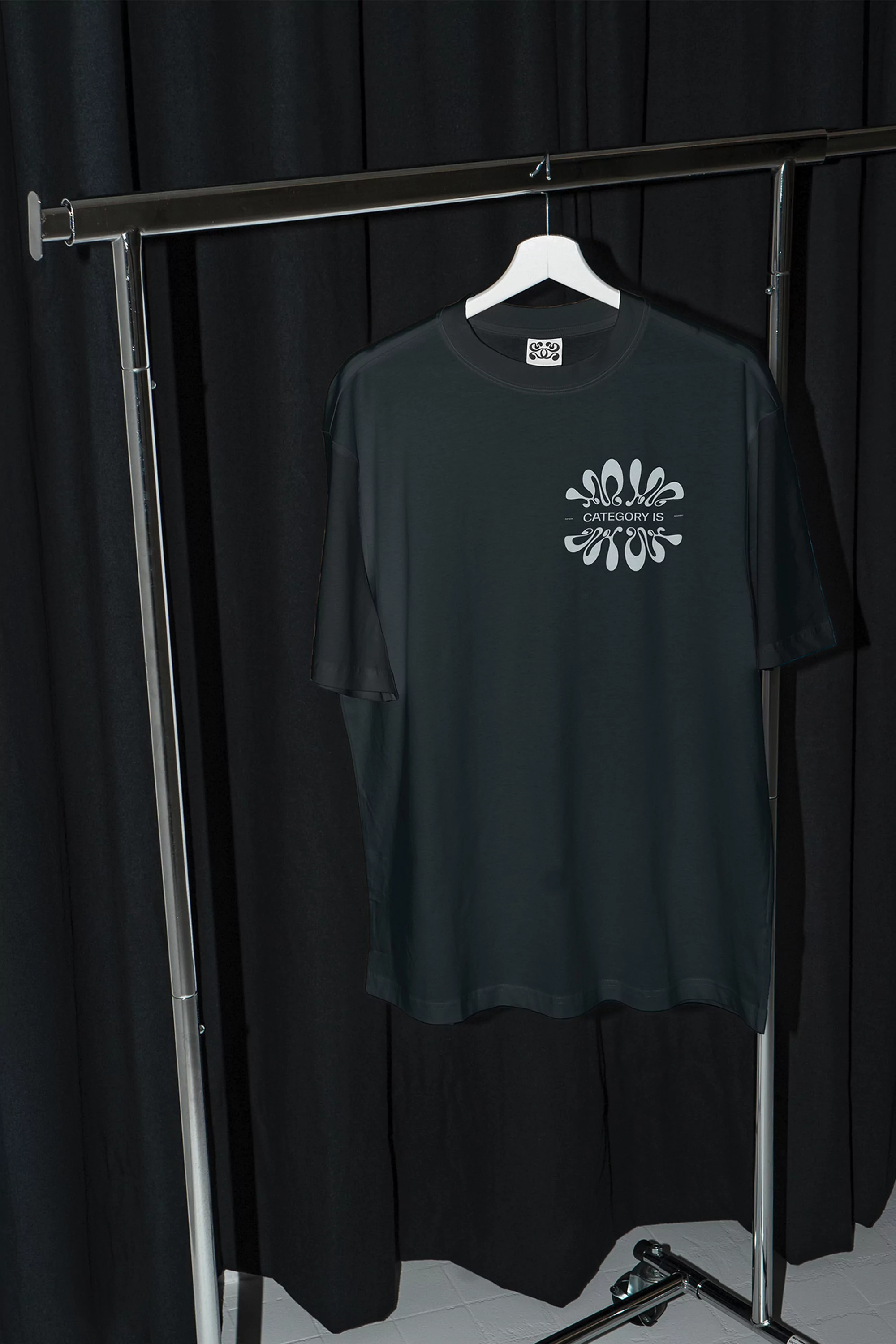 Black t-shirt with white graphic design hanging on a white hanger displayed on a metal clothing rack against a black background.