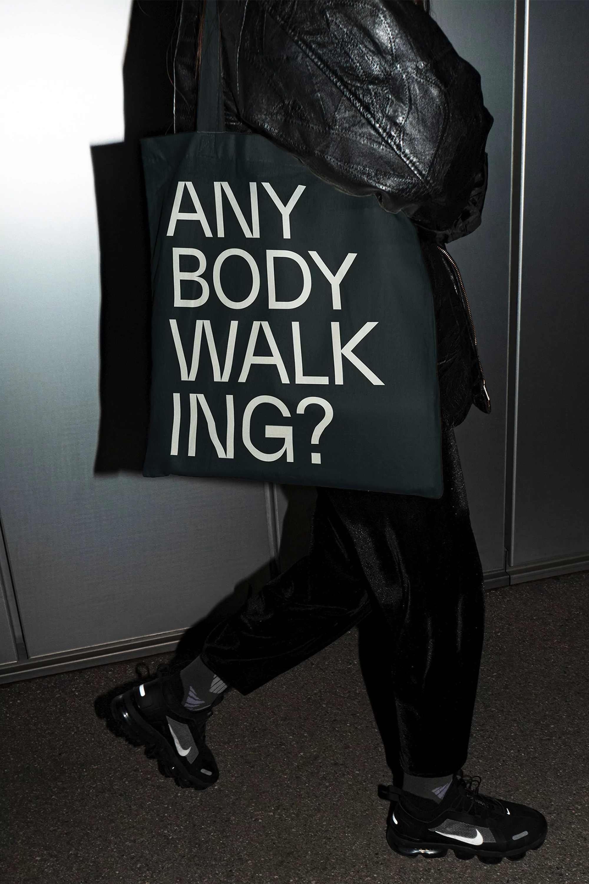 Person wearing black apparel holding a dark tote bag with bold typographic text against a neutral metal panel wall