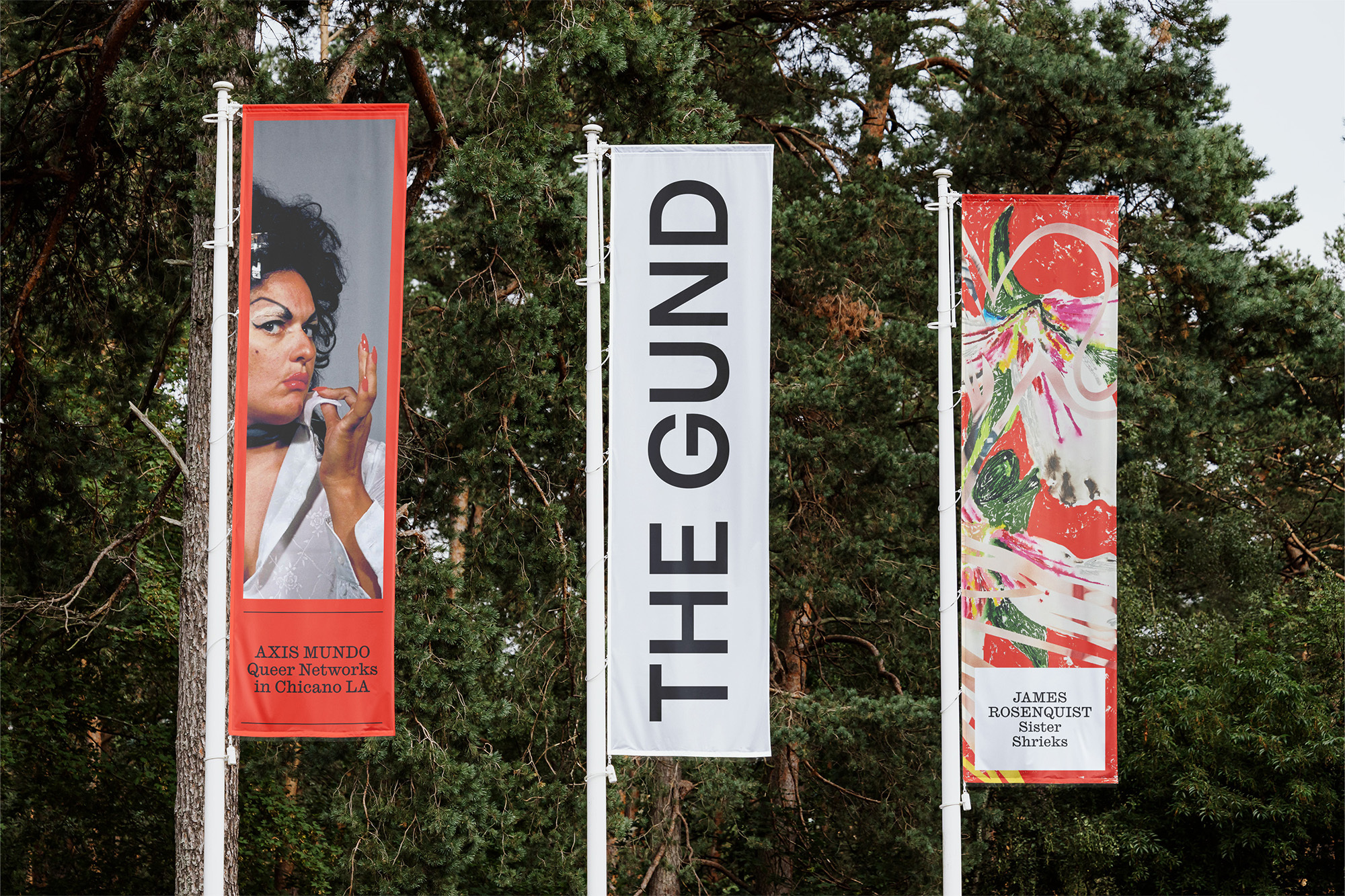Three vertical advertising banners with images and text are displayed outdoors, framed by green trees and set against a daytime sky.