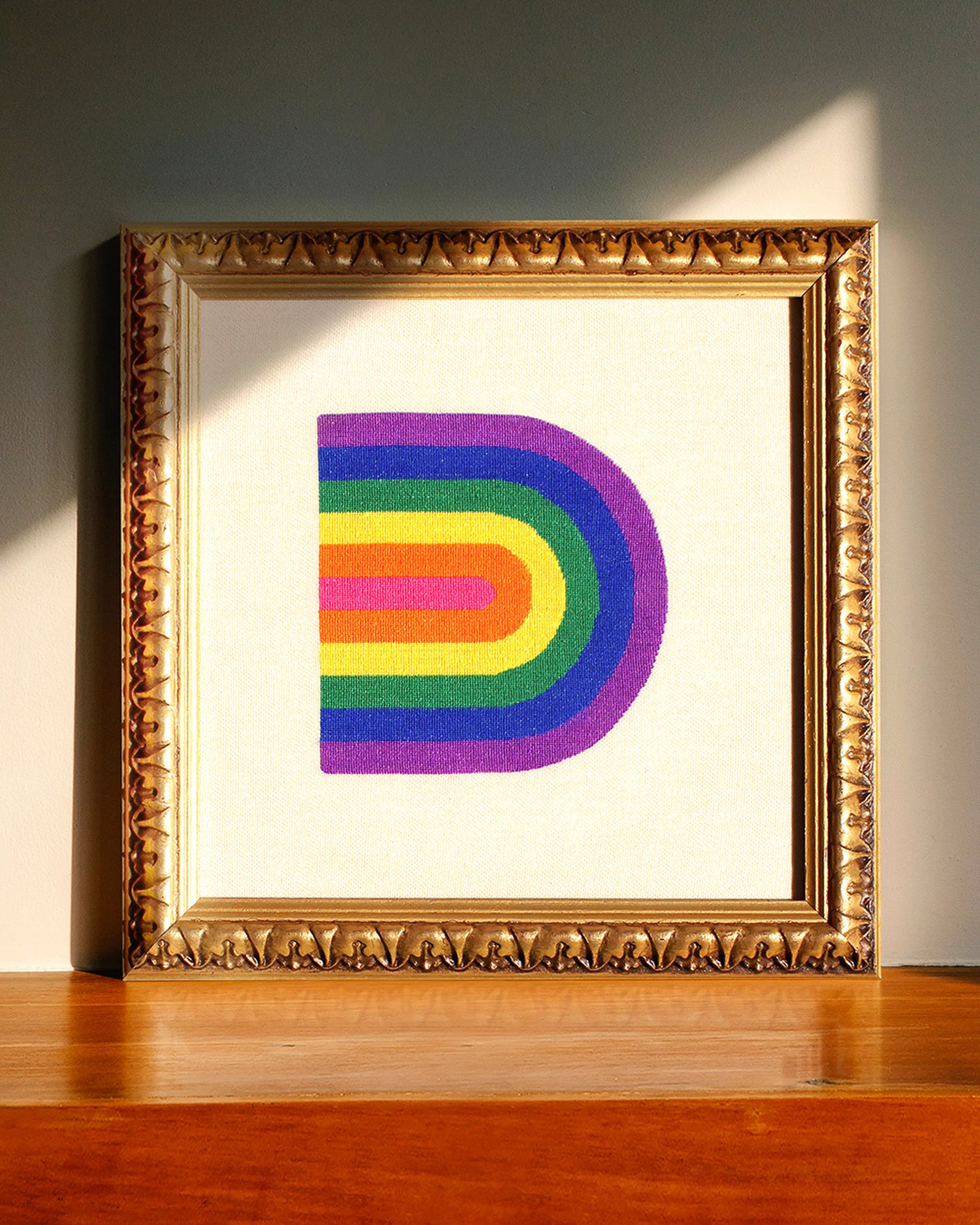 Framed art features a colorful rainbow design on canvas, set against a neutral wall and a warm wood surface, illuminated by natural light.