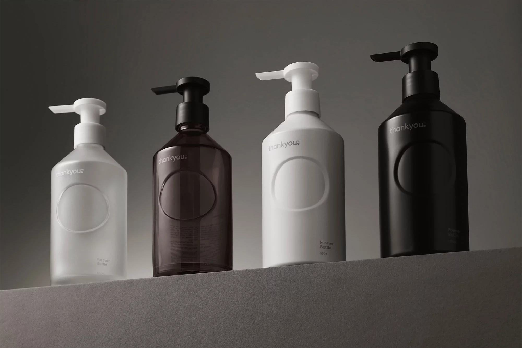 Four Thankyou Forever Bottles in white, brown, and black colors with pump dispensers, arranged on a neutral gray surface against a gray background.