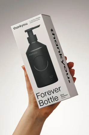 Hand holds Thankyou Forever Bottle in packaging: matte black refillable bottle with pump dispenser shown in white and black box.