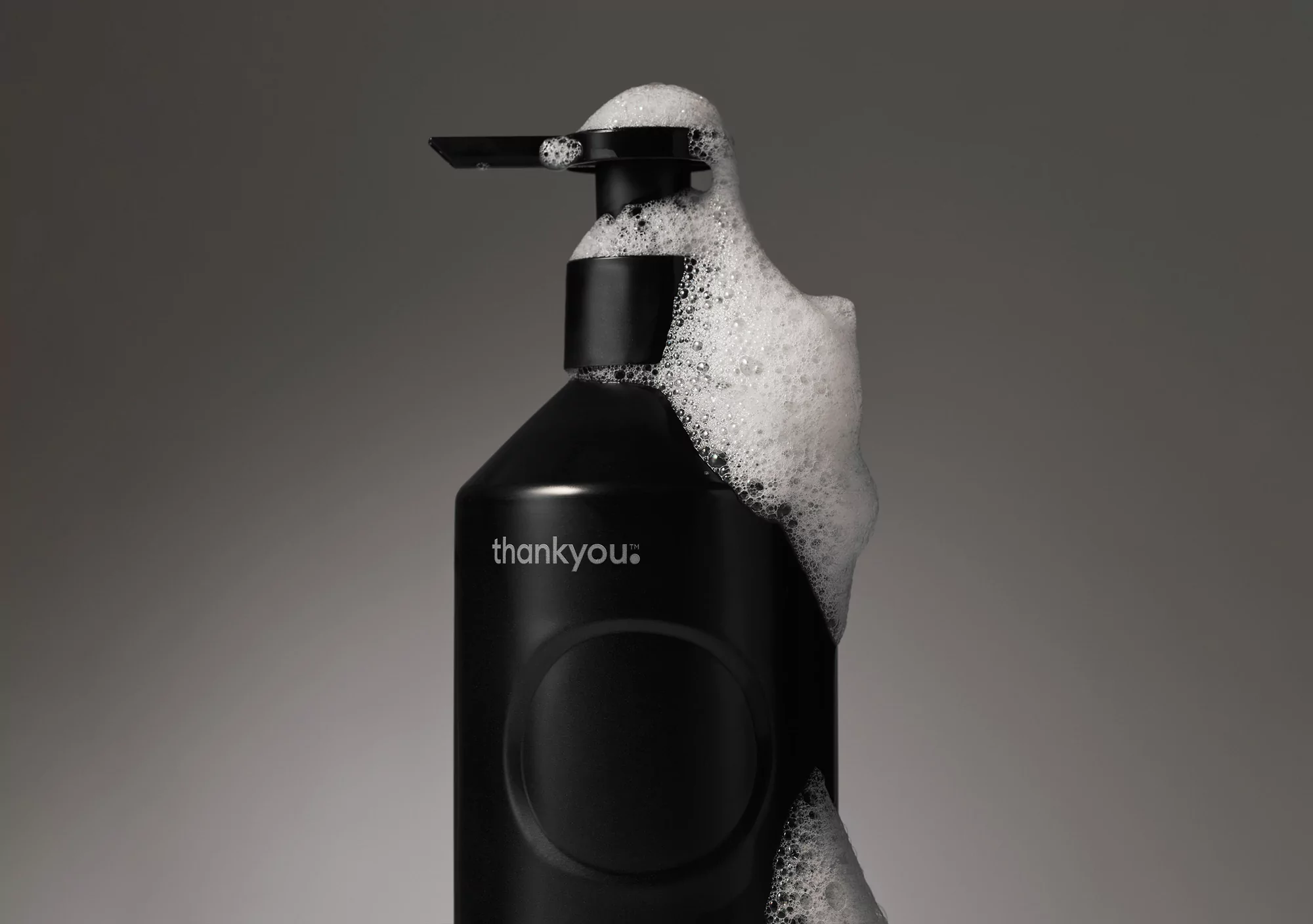 Black dispenser bottle with pump surrounded by white foam, set against a grey gradient backdrop, showcasing beauty and hygiene product.