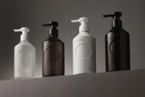 Four pump bottles in translucent frosted, dark brown, solid white, and solid black arranged in a row on a grey ledge.