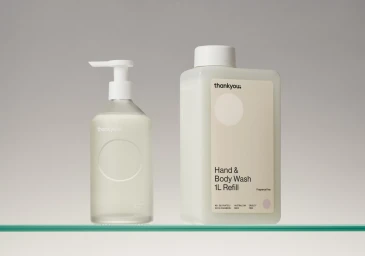 Two Thankyou brand refillable body wash bottles standing on a transparent glass shelf against a neutral grey seamless background
